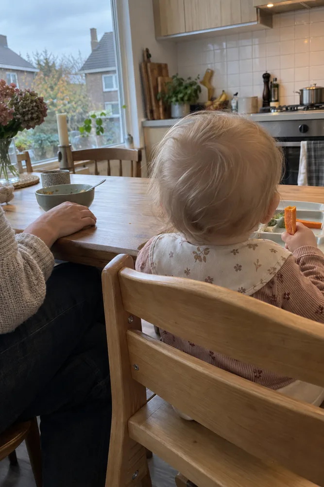 Een baby van achteren gezien, rechtop in een houten kinderstoel, met een volwassene ernaast